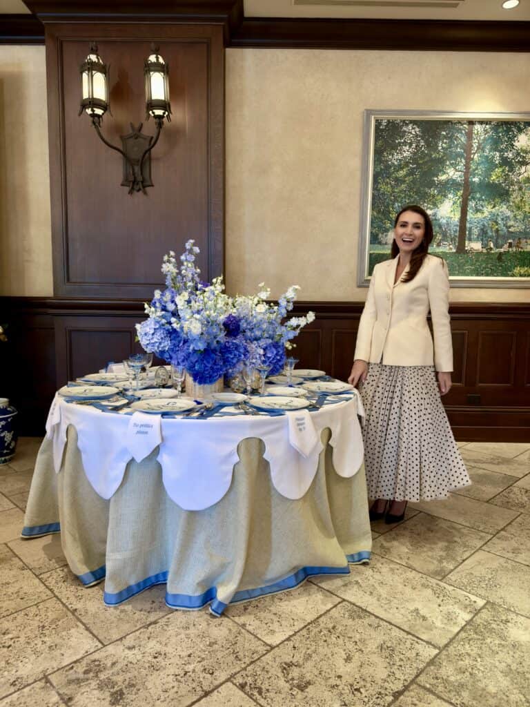 Kimberly Schlegel Whitman wearing Dior Bar Jacket at a blue and white table for Kappa Tablescapes. Table inspired by blue ribbons and bows with Please Leave By 9 napkins.